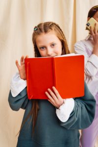 A young girl with braided hair reads a bright red book indoors, capturing moments of curiosity and learning.